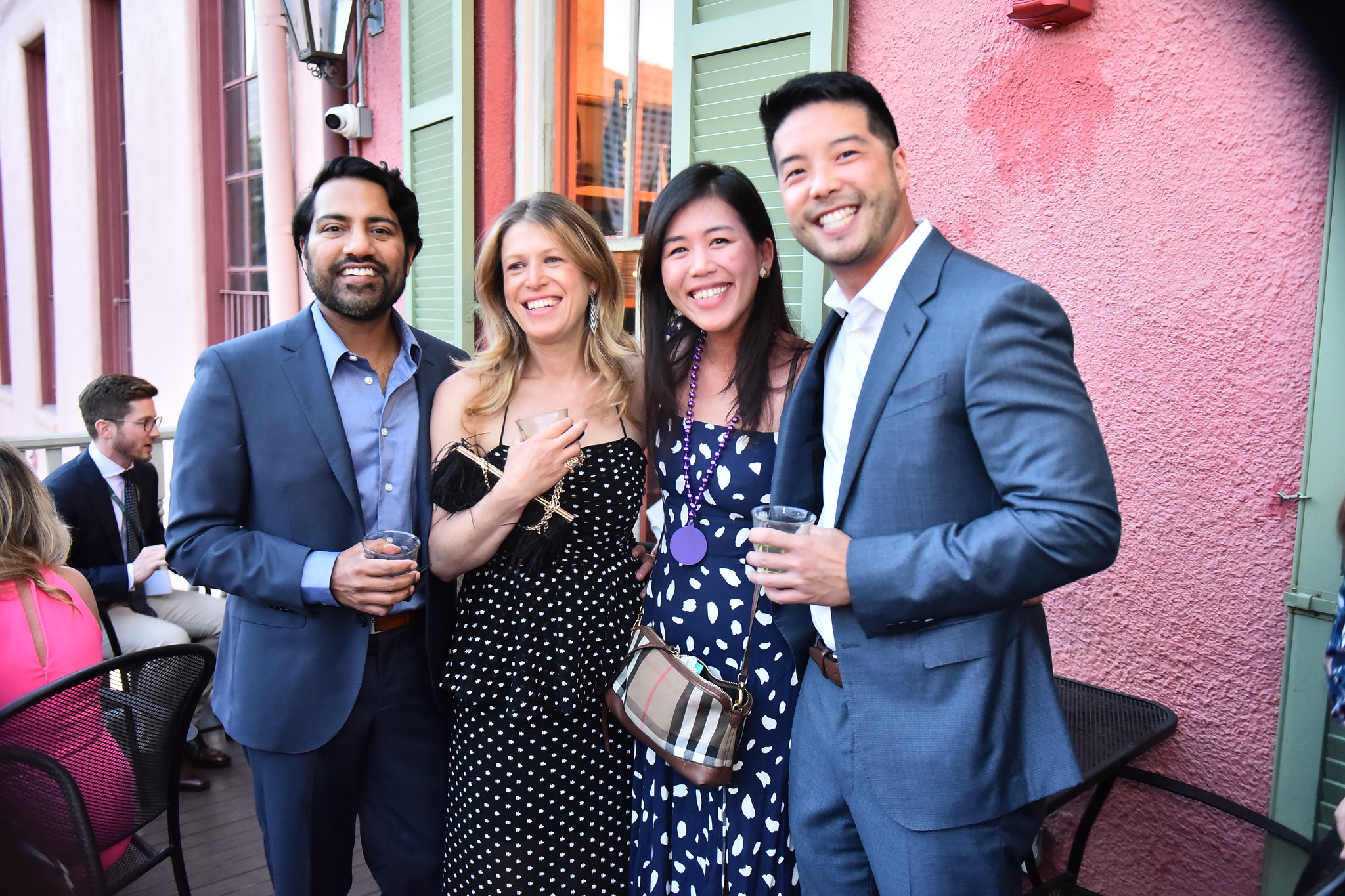 Four smiling people, two men and two women, holding drinks on an outdoor patio. Pink building in background.