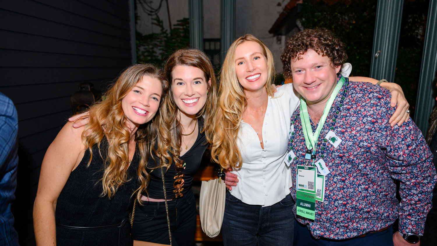 Four smiling friends, three women and a man, pose closely at a nighttime event, green lanyard visible.