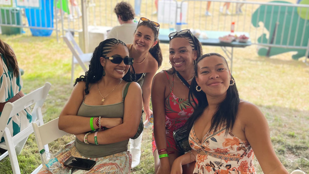 Four smiling women at an outdoor festival, two seated and two standing.