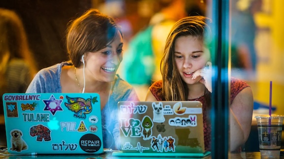 Two smiling young women look at laptops, seen through a window.