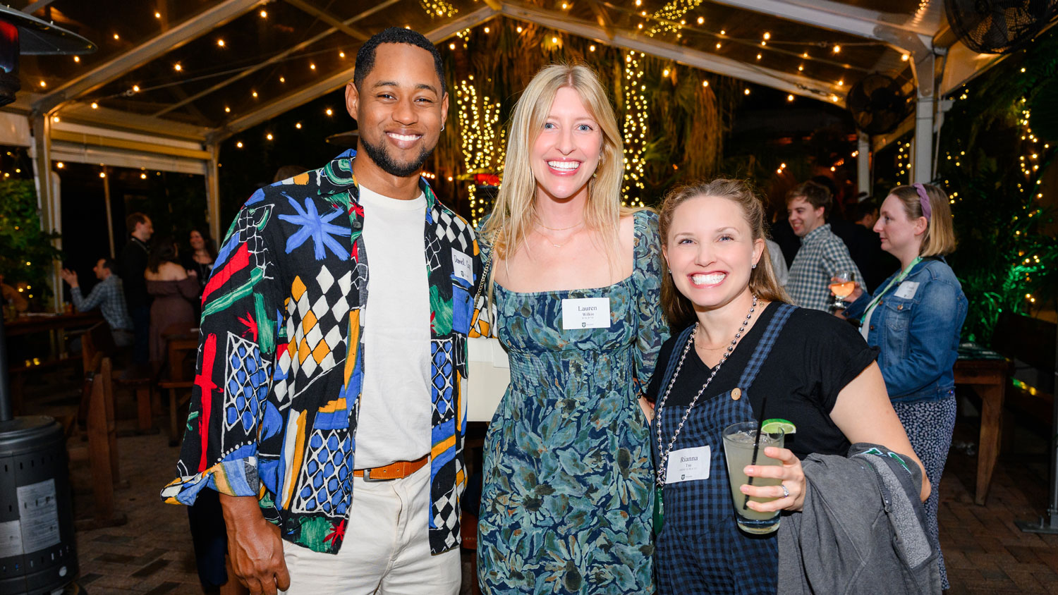 Three smiling people pose under string lights at an outdoor evening event.