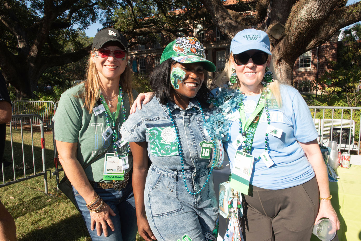 Three smiling women in green and blue outfits with face paint and lanyards at an outdoor event.
