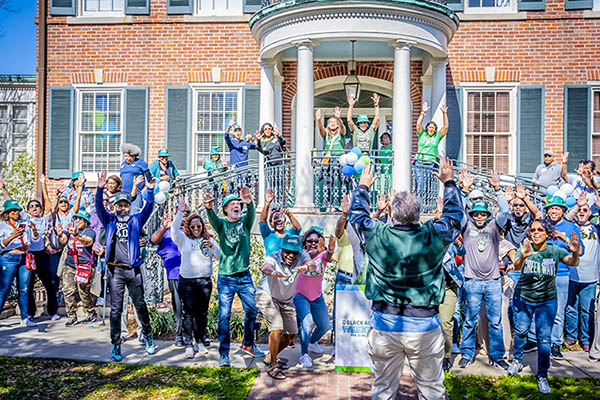 Large group of diverse people cheering with arms raised in front of a brick building.