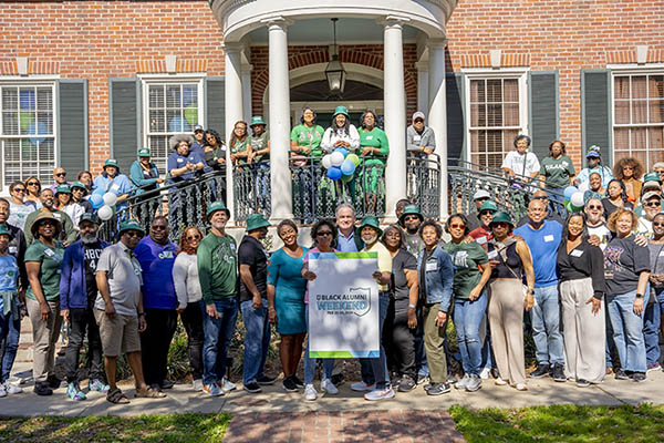 Group in green and blue shirts with "Community Health" sign in front of a brick house.