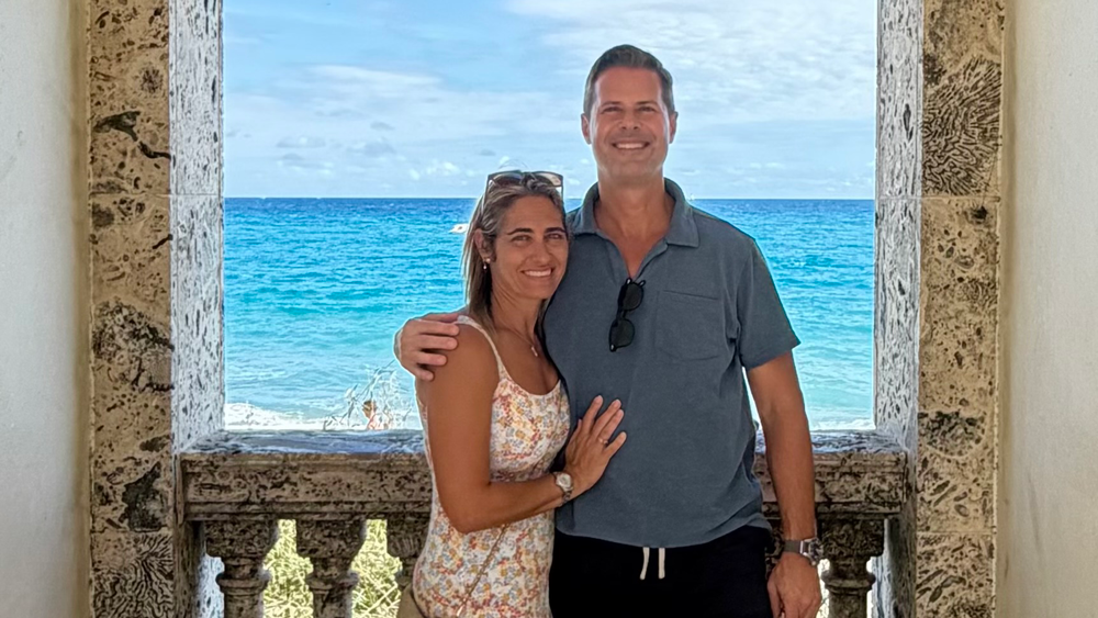 Smiling couple on a stone balcony with a bright blue ocean view.