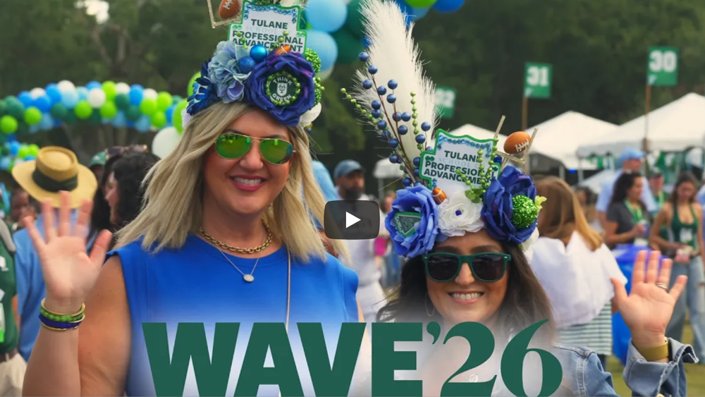 Two women in festive blue and green hats and sunglasses wave, smiling outdoors.