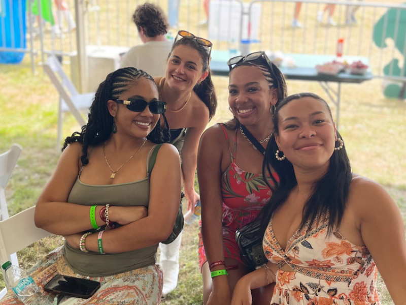 Four smiling women at an outdoor festival, two seated and two standing.